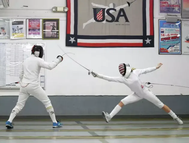 Two fencers in white gear and masks are in a duel inside a gym, with the one on the right performing a lunge.