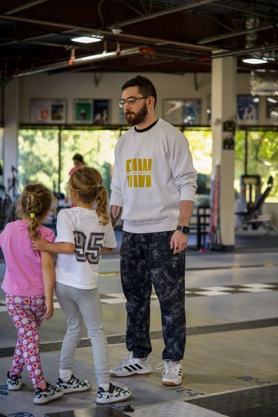 A man in a sweatshirt with two young girls in a gym. Man is speaking to girls.