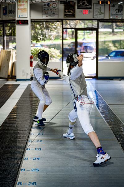 Two fencers in protective gear dueling on a strip. One lunges, the other parries. Indoors, fencing club.