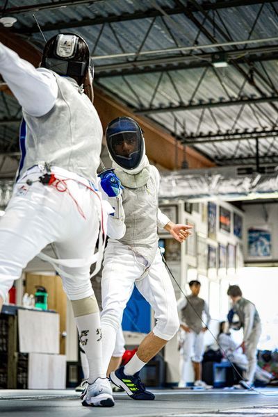 Two fencers in white gear, masked, lunging at each other. Fencing in a gym.