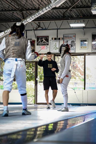 Two fencers and coach on a fencing strip, coach gesturing with a foil.