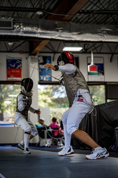 Two fencers in protective gear compete, one lunging forward with a foil. Indoor setting, with spectators visible.