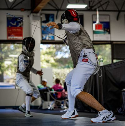 Two fencers in protective gear spar on an indoor strip, one lunging forward with an extended foil while the other defends.