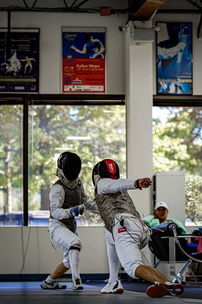 Two fencers in protective gear, fencing in a gym. One lunges forward, arm extended.