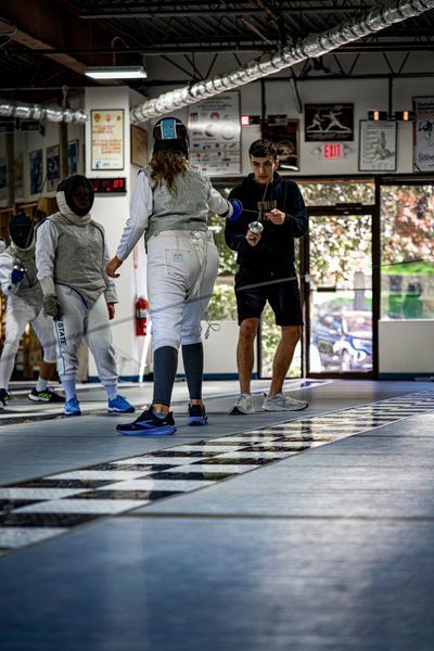 Fencers in a gym, practicing with coach. One person in focus holding a sword.