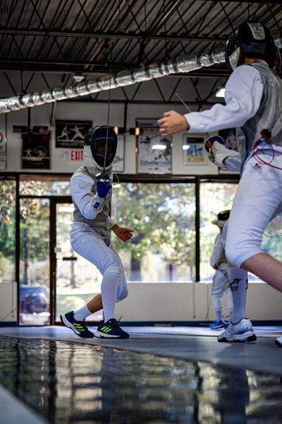 Two fencers in protective gear on a reflective floor, lunging and dueling in a brightly lit space.