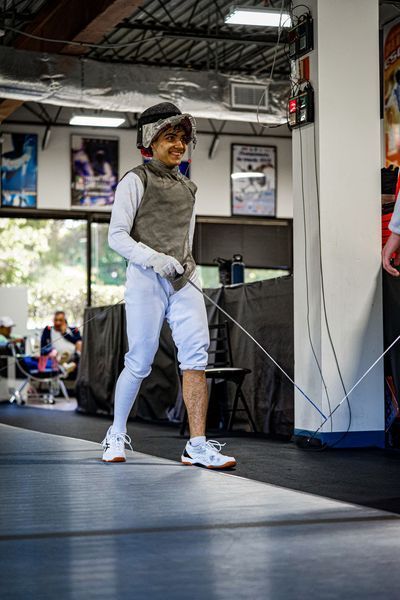 A person in fencing gear walks on a strip indoors, smiling.