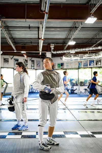 Fencer in white uniform stands in a gym, holding a foil. Other fencers are in the background.