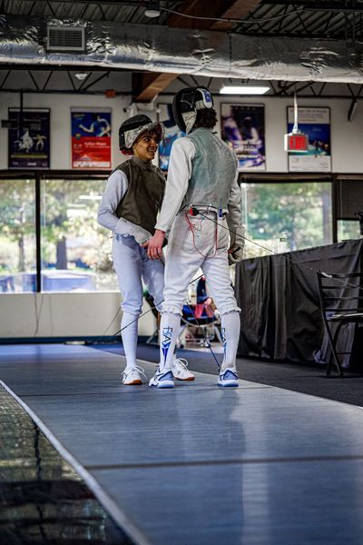 Two fencers in protective gear on a fencing strip, face each other, likely preparing to bout.