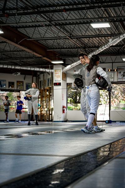 People in fencing gear on a blue strip in a gym. One holds a weapon, others watch.