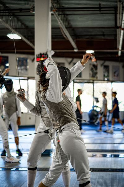 Two fencers in white uniforms, dueling. One lunges forward with foil, the other deflects, indoors.