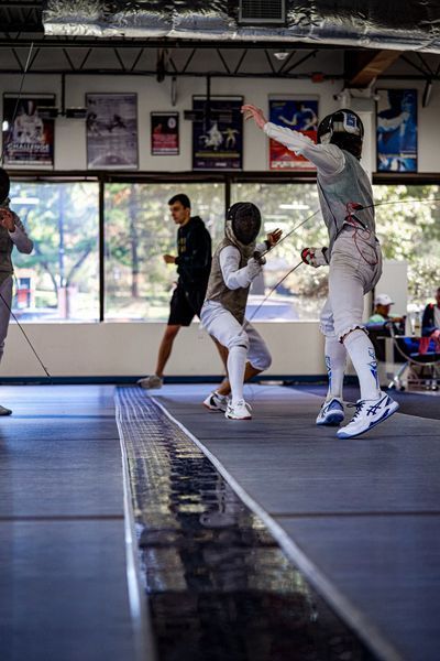 Fencers in action on a strip, indoor fencing gym setting. One raises arm, another lunges, others watch.