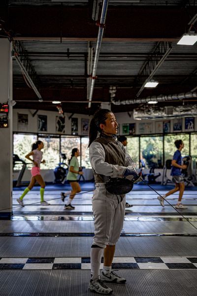 Fencer in uniform looks on, holding a mask, with others fencing and running in the background, in a gymnasium.