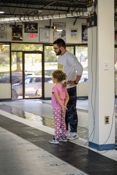 Man and young girl watch something in a gym. Man is looking forward, hands on hips. Girl stands with hands behind her back.