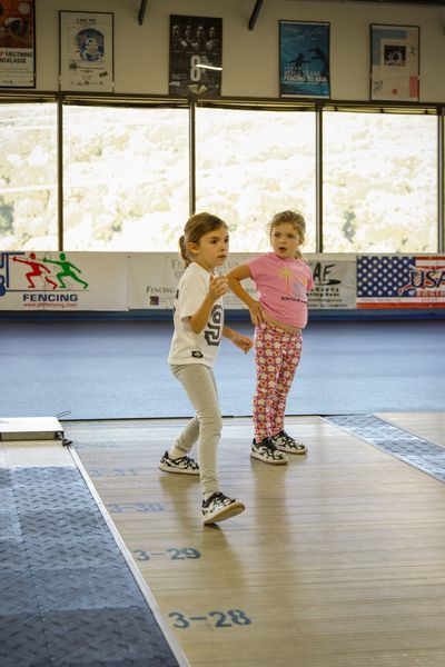 Two young children in a fencing gym, one lunging, the other watching.
