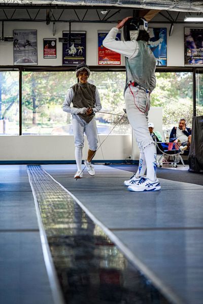 Two fencers in a fencing studio, one adjusting helmet, the other facing forward.