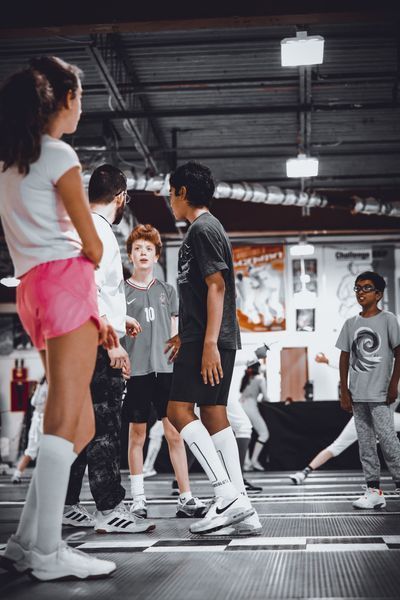 People at a fencing training gym. One person in pink shorts stands, others in athletic wear.