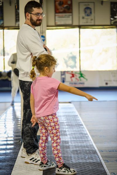 A child in pink points on a fencing strip, next to a man. Light fills the room.