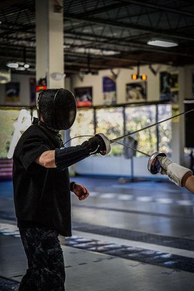 Two fencers in protective gear, fencing in a gymnasium with a metallic floor. One in black attacks.