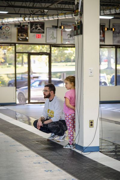 Man and child watching; man crouches, child stands beside a pillar in a sports facility; windows and cars visible.