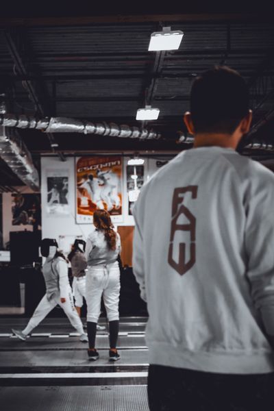 A group of young fencers in white gear standing in a line at a fencing club, holding masks in front of a tournament chart.