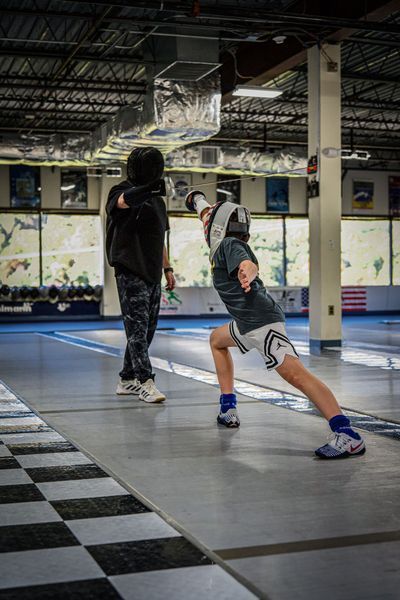 Fencer in a white helmet lunges toward opponent, both in fencing gear, on a marked gym floor.