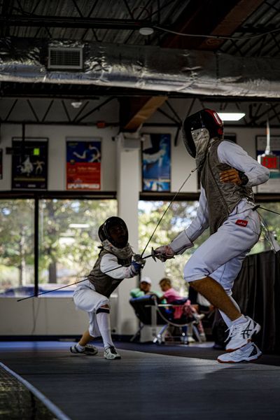 Two fencers in a brightly lit indoor space, lunging with foils. One is smaller. White and brown protective gear.