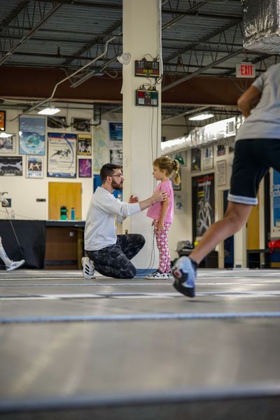 A coach kneels, guiding a child in pink pajamas in a gym. A person in shorts runs nearby.
