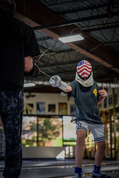 Fencer in an American flag mask lunges toward opponent. Indoors, foil raised. Opponent partially visible.