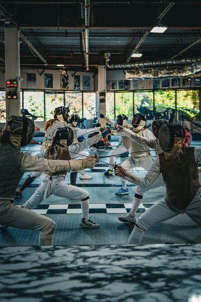 Fencers in protective gear practicing in a studio with a checkered floor, locked in a forward lunge with foils extended.