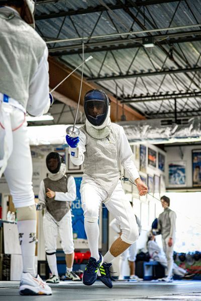 Fencer in white uniform lunges forward with foil, another fencer in background. Indoor setting, bright.