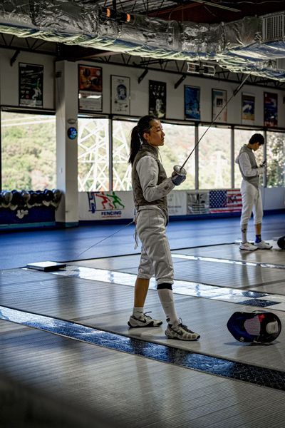 Fencer in white uniform with foil, standing on a blue mat. Another fencer in background. Indoor setting.