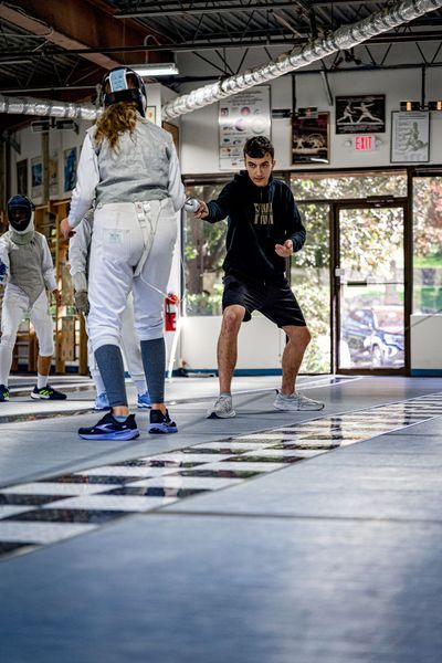 Fencing students practice in a gym. A person in black lunges with a foil. Others watch.