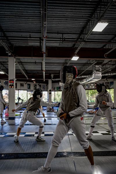 Fencers in protective gear practice in a gymnasium. One in focus, others in background, on a black and white checkered floor.
