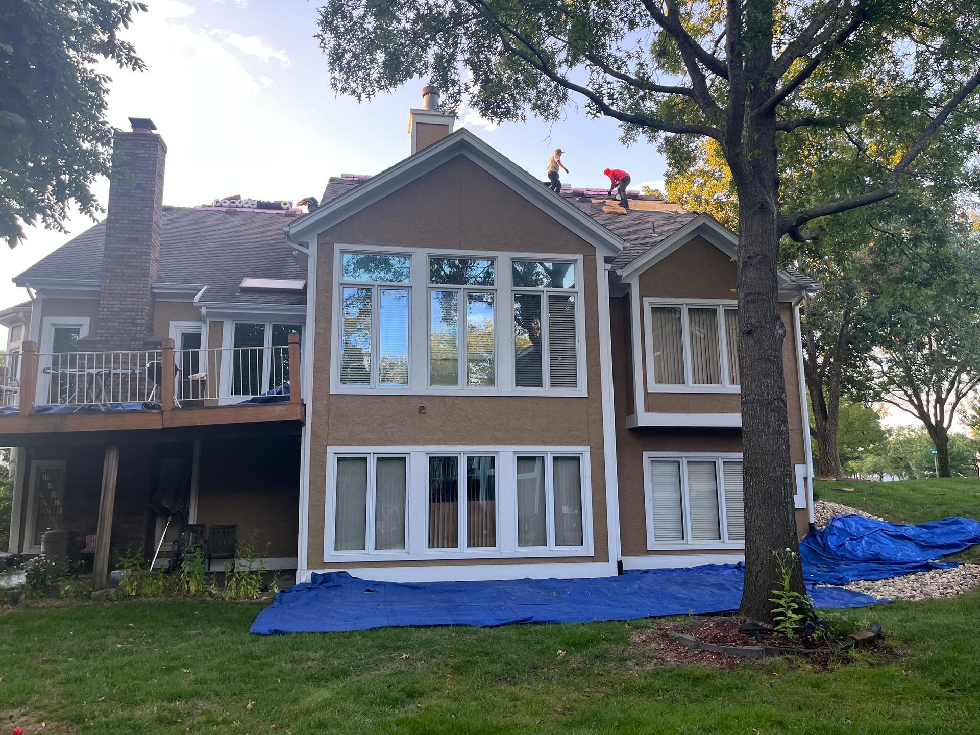 House exterior with roofers on roof, brown stucco siding, blue tarp on grass.