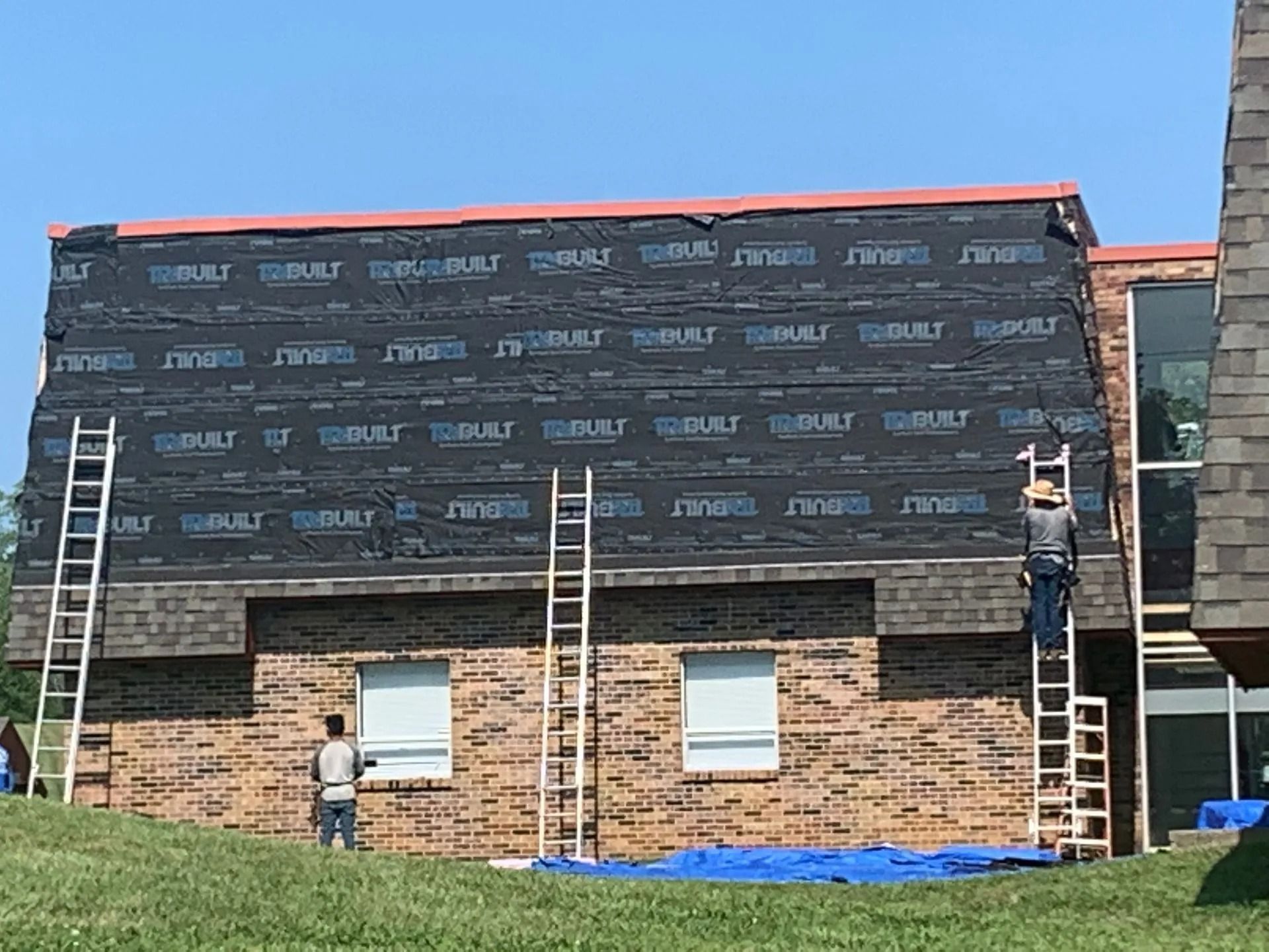 Construction workers installing roofing on a brick building, ladders propped against the side, blue tarp below.