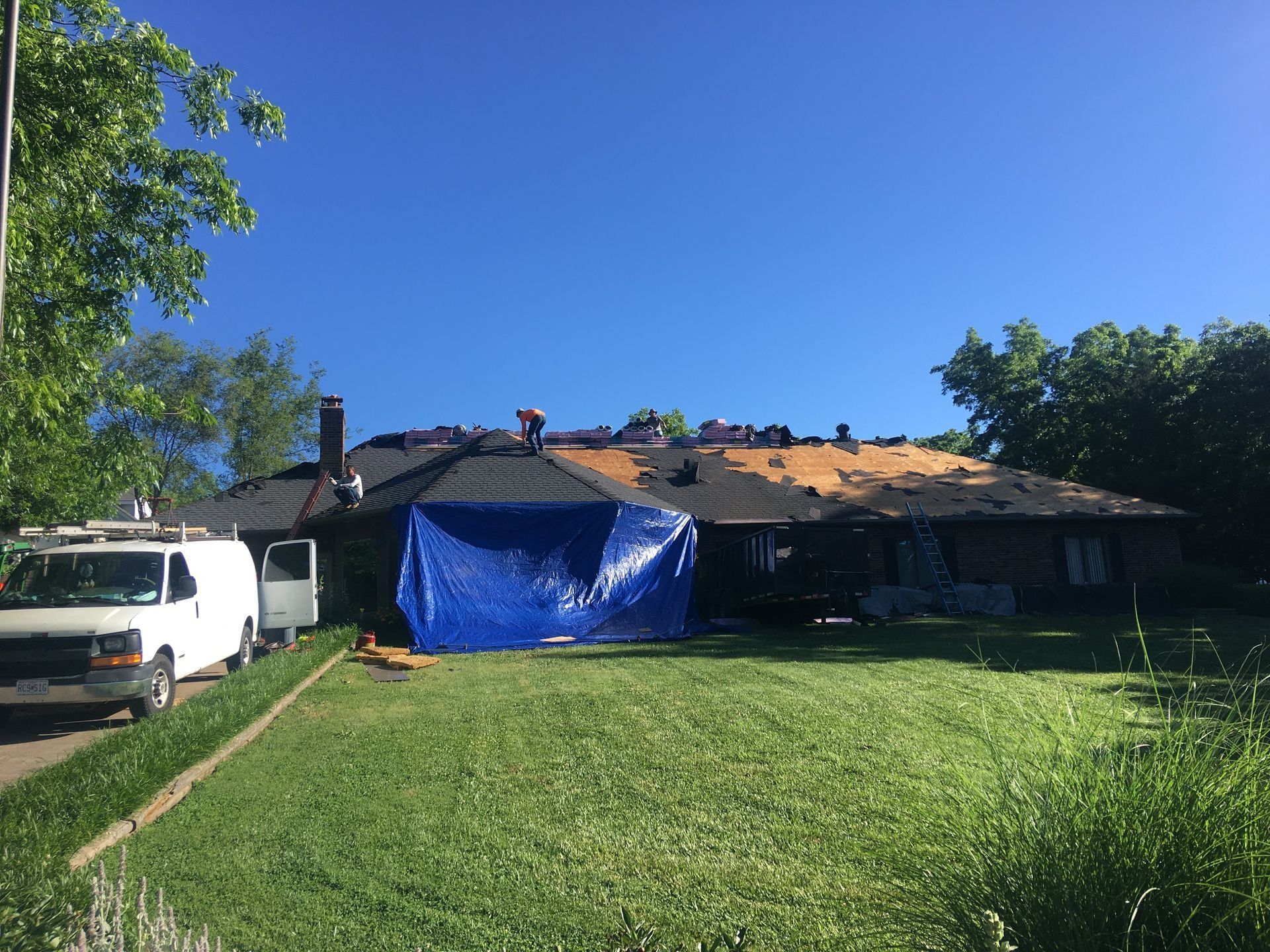 Roofers working on a house with a blue tarp covering a section; white van parked on the lawn.