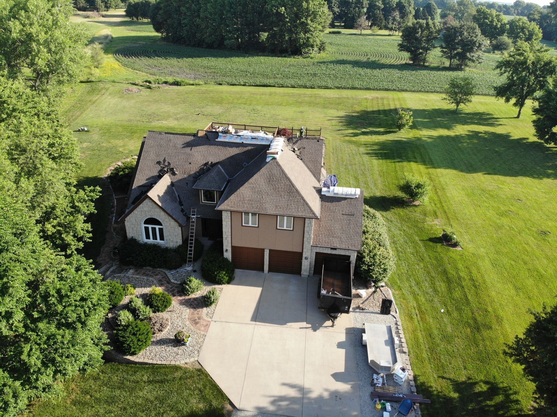Aerial view of a brown-roofed house with two-car garage, surrounded by green fields and trees on a sunny day.