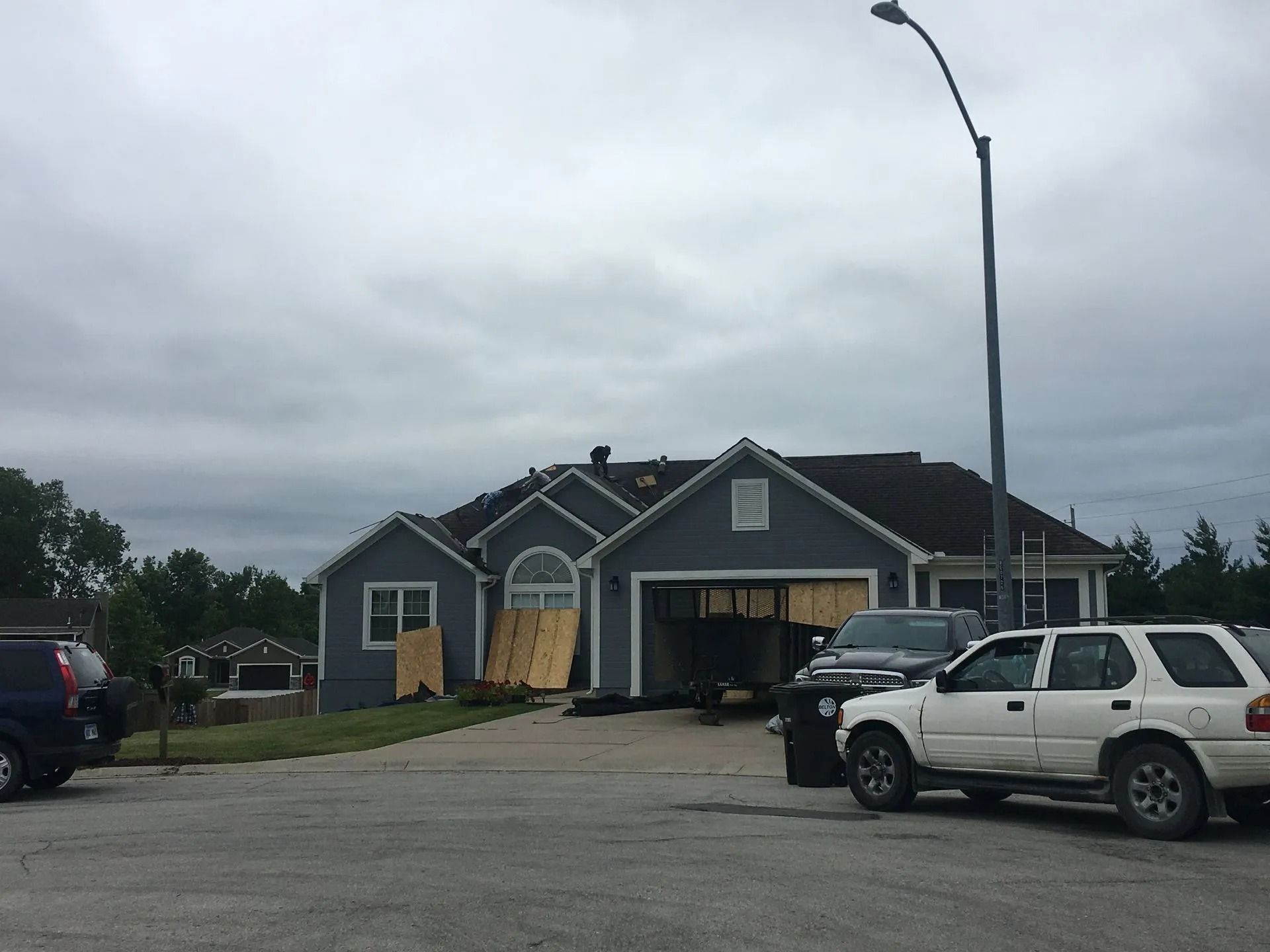 Damaged house with boarded-up garage door after storm. Cars parked in driveway. Gray sky overhead.