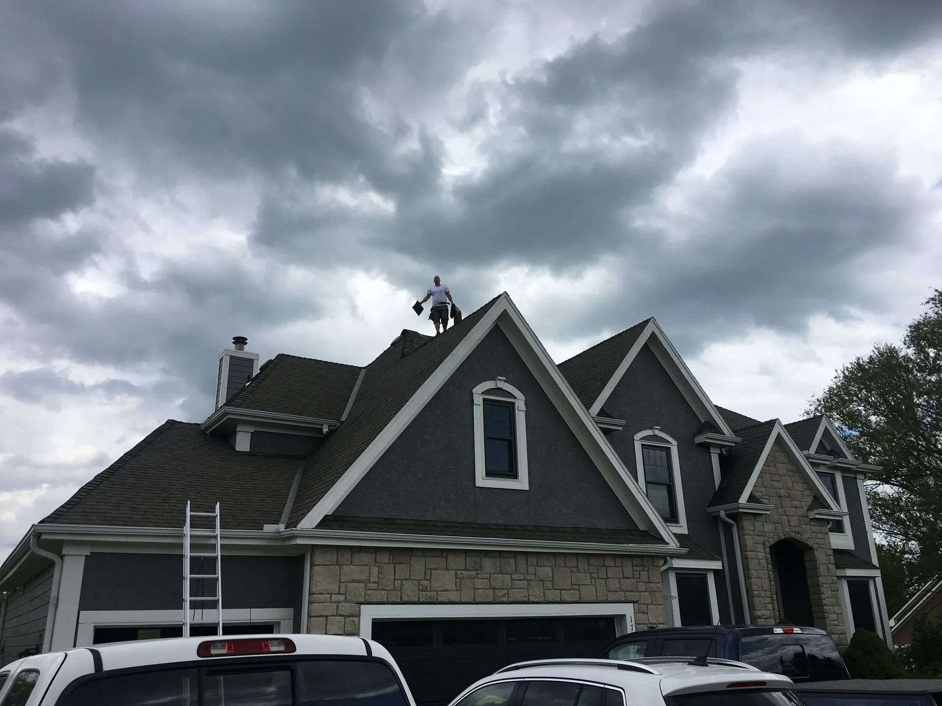 Person on roof of house with dark gray siding, under cloudy sky. Ladder against the side.