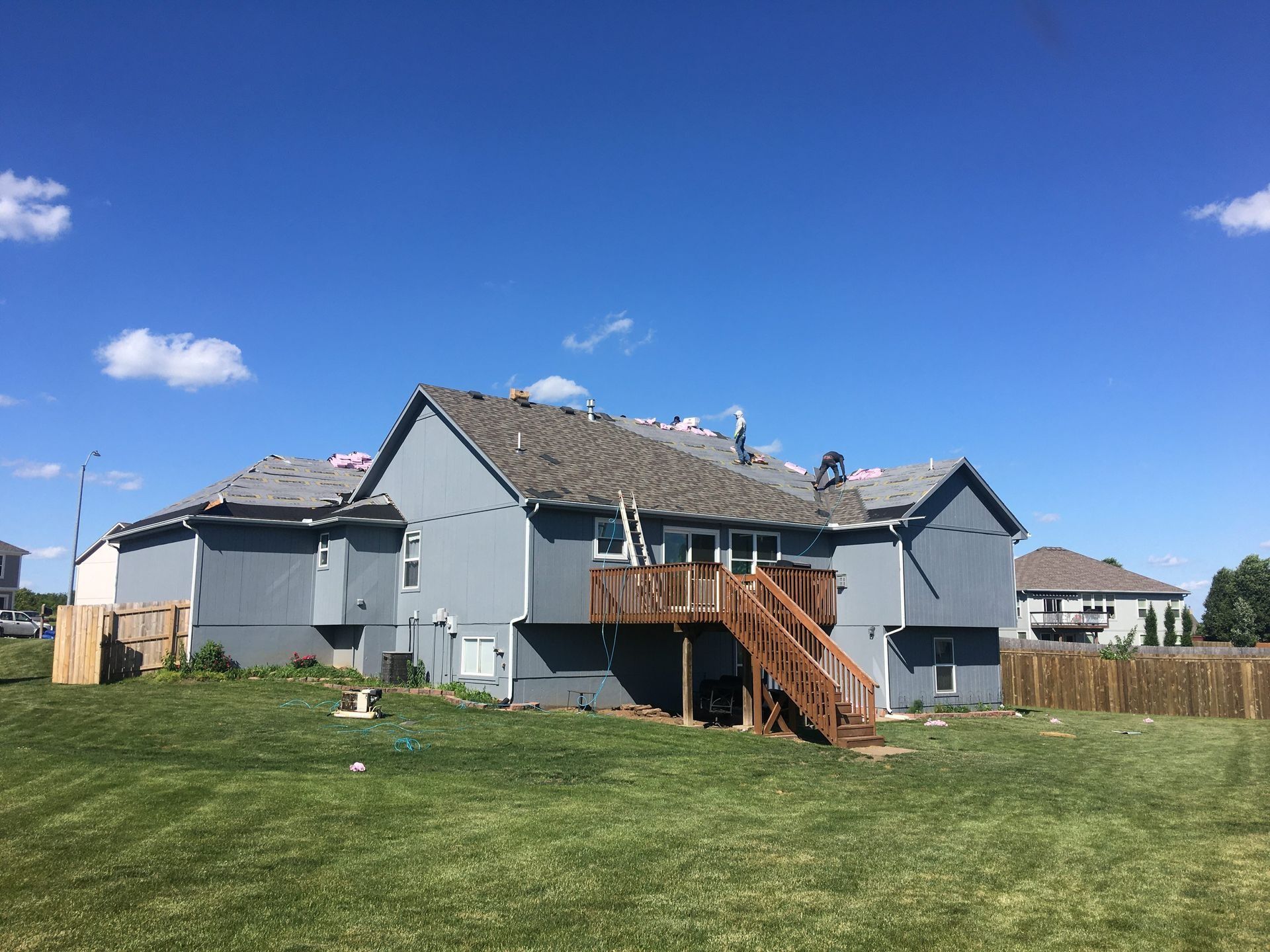Backyard of a blue house with wooden deck, grey roof, and green lawn under a blue sky.