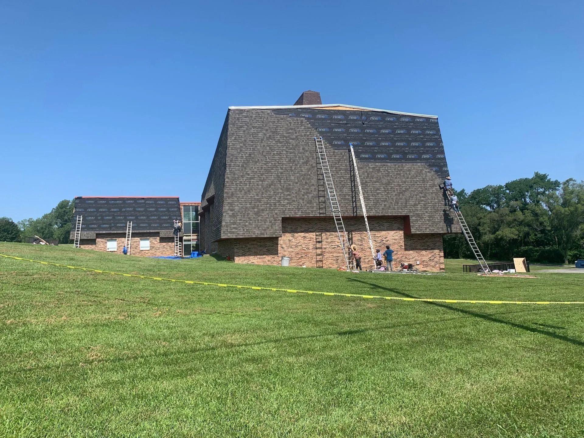 Roofers on ladders working on a multi-story brick building with a partially completed shingle roof, green lawn, and blue sky.