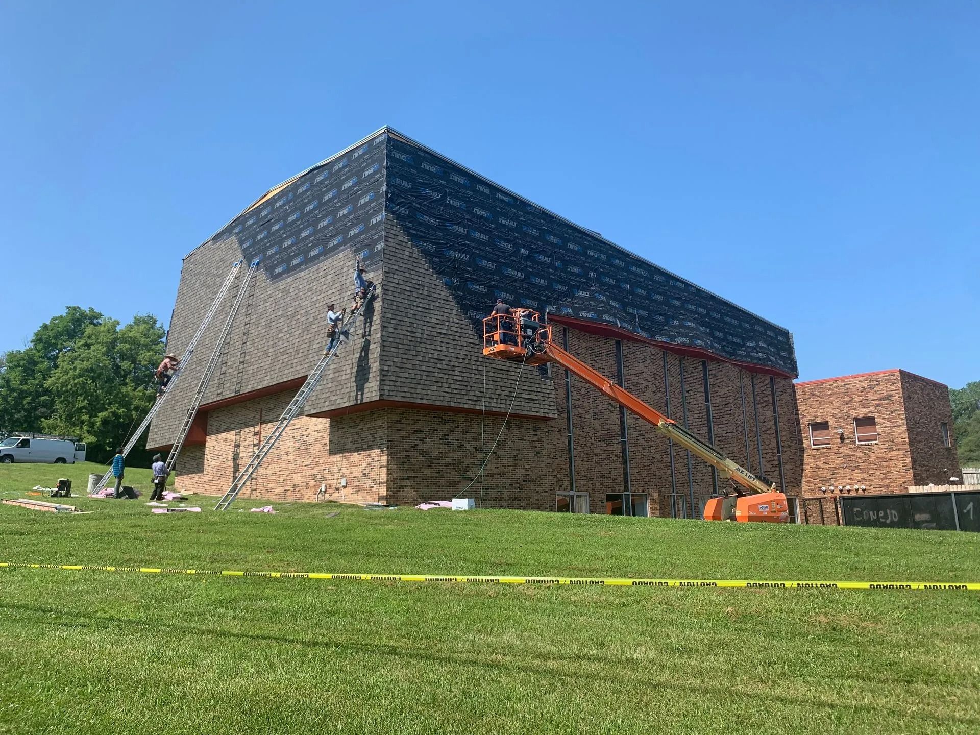Building construction: exterior partially covered with dark roofing and brick, lift in use, workers present.
