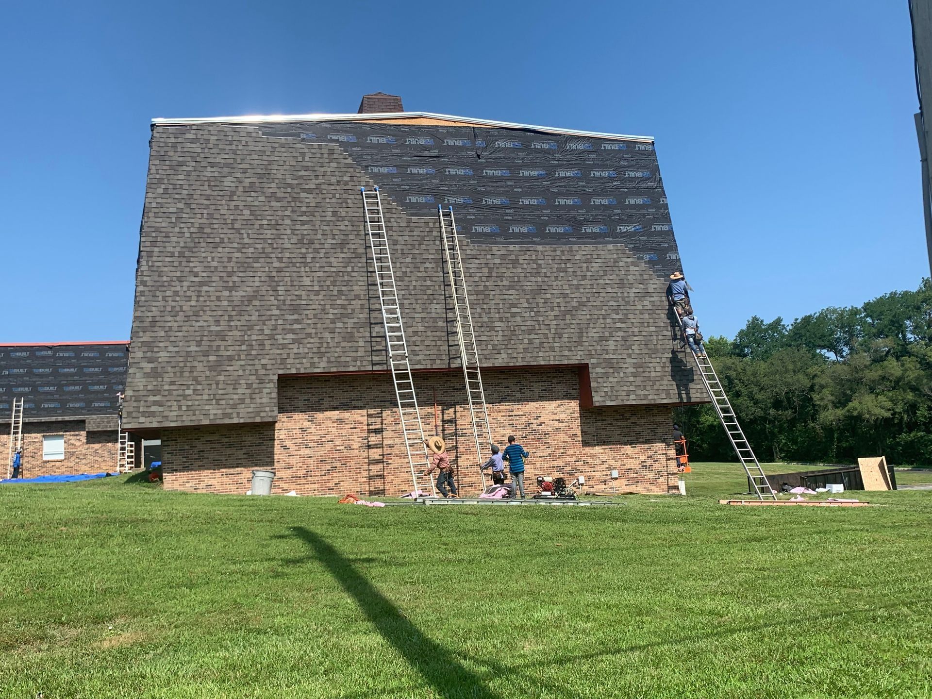 Construction workers on ladders roofing a brick building on a sunny day.