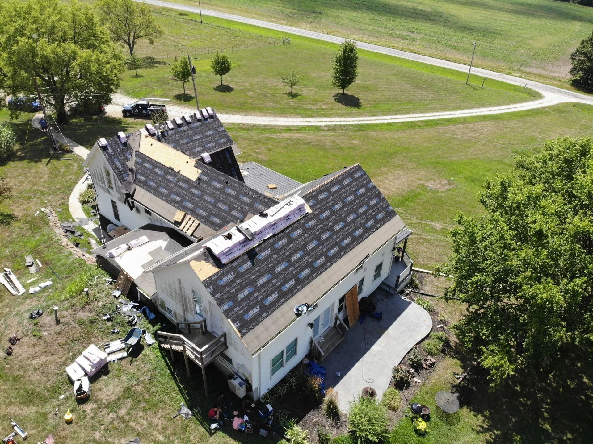 Aerial view of a house with a partially replaced roof; debris on the ground. Green grass surrounds the house, and a road is in the background.