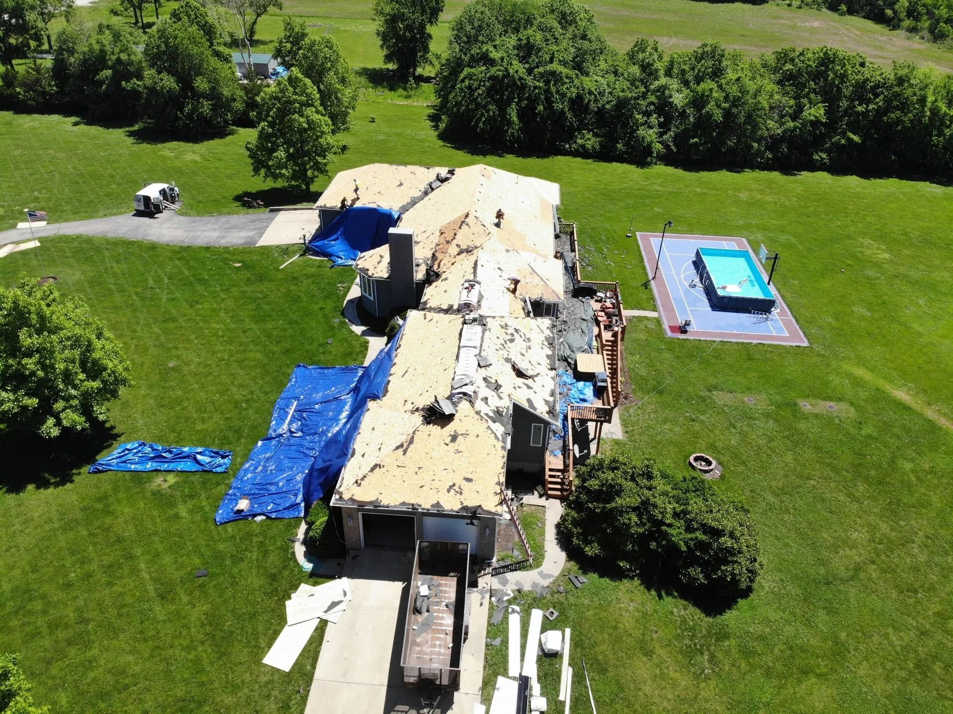 Aerial view of a house with roof damage, covered in blue tarps, on green grass. There is a pool in the yard.