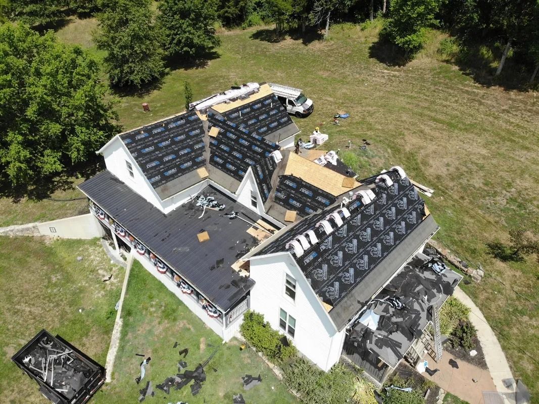Overhead view of a house with partially completed roof replacement, surrounded by green grass and trees.