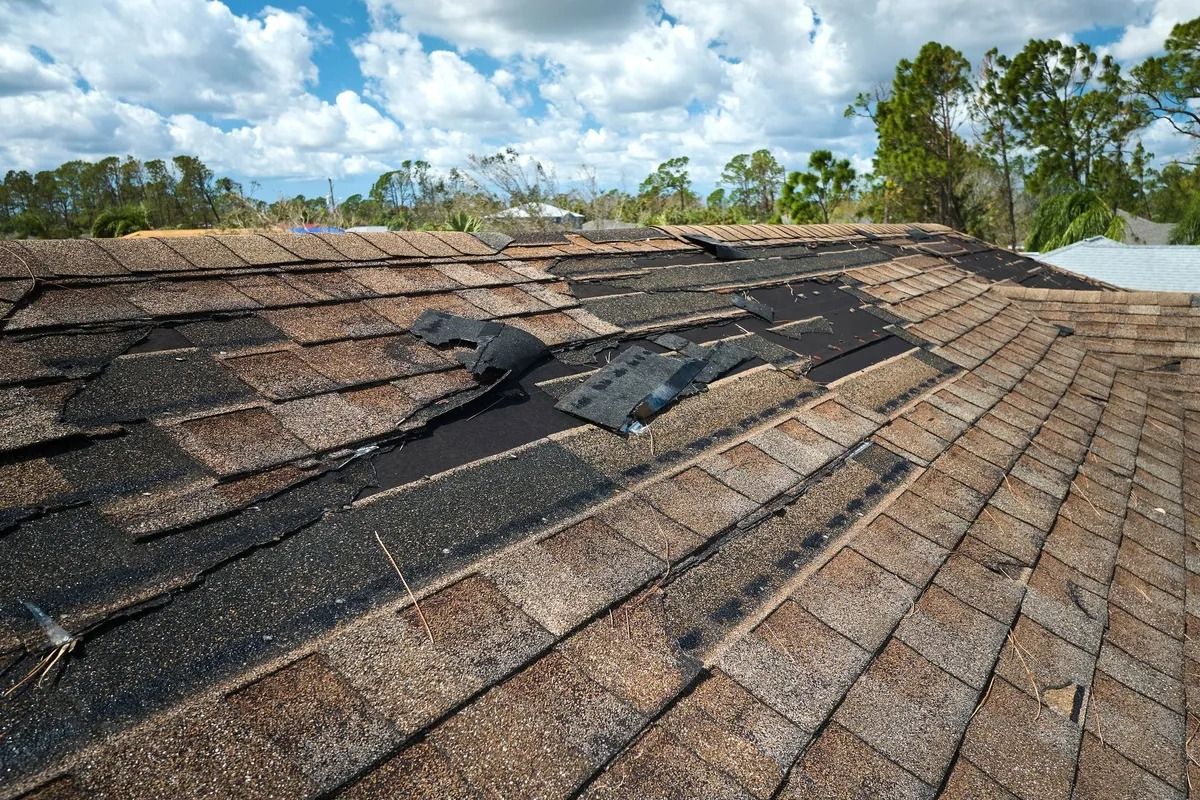 Damaged asphalt shingle roof, with missing and torn areas, under a partly cloudy sky.