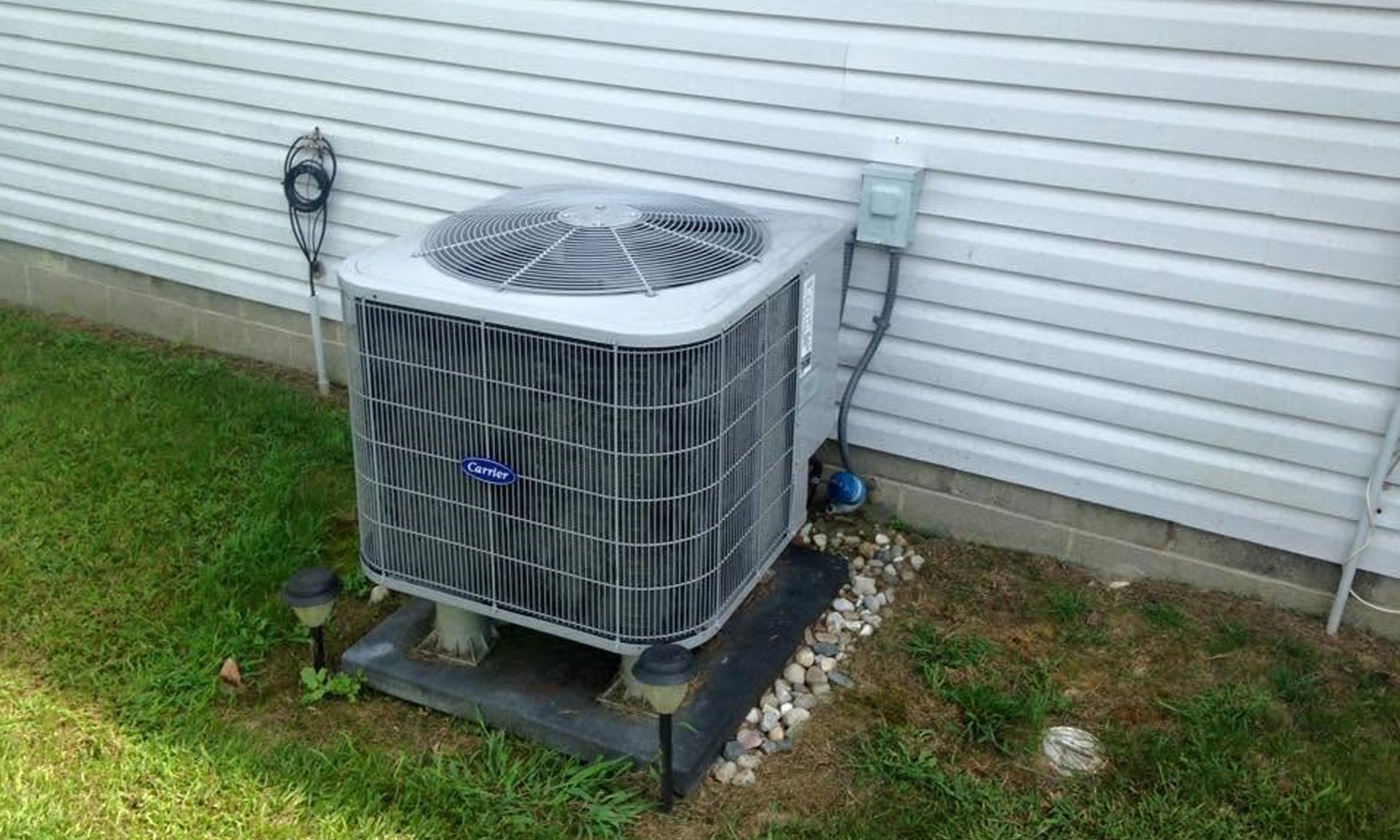 Outdoor air conditioning unit beside a house wall on a metal stand, with grass and gravel around it