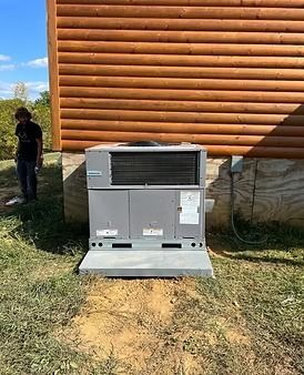 Outdoor HVAC unit beside a log building, with a person standing to the left on a grassy patch.